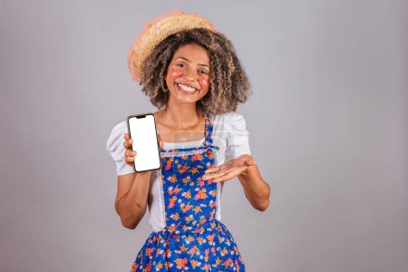 Photo for Young black Brazilian woman, with country clothes, dressed for Festa Junina. holding smartphone with white screen for ads, mobile apps. - Royalty Free Image