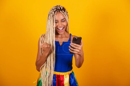 Photo for Beautiful black, brazilian woman with braids, wearing clothes for carnival. Holding smartphone. cheering. - Royalty Free Image