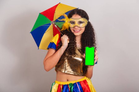 Téléchargez les photos : Jeune adolescente, brésilienne, avec des vêtements frevo, carnaval. avec parapluie frevo. tenant smartphone avec écran chromé vert. - en image libre de droit