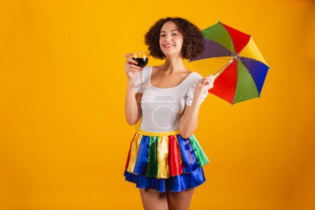 Téléchargez les photos : Belle femme brésilienne, vêtue de vêtements de carnaval, jupe colorée et chemise blanche. parapluie frevo, tenant un verre de vin. - en image libre de droit