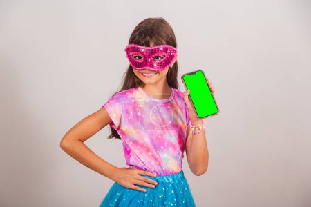Photo for Beautiful Brazilian girl, child, dressed for carnival in Brazil. hands on waist with smartphone with green Chroma screen. - Royalty Free Image