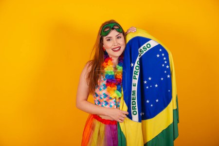 Photo for Brazilian redhead, in carnival clothes, with a flower necklace around her neck, holding a Brazilian flag. - Royalty Free Image