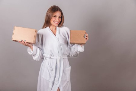 Photo for Brazilian blonde woman showing wonderful hair. dressed in a robe. holding two cardboard boxes with cosmetics. - Royalty Free Image