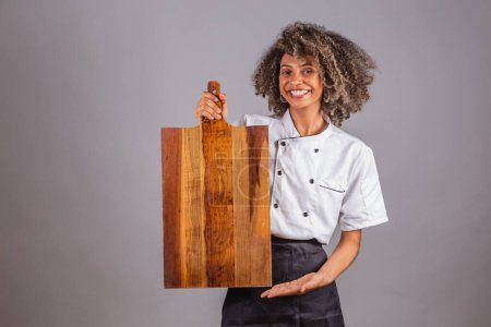 Photo for Young black Brazilian woman, cook, masterchef, wearing restaurant uniform. holding wooden board for texts and advertisements, presenting. - Royalty Free Image