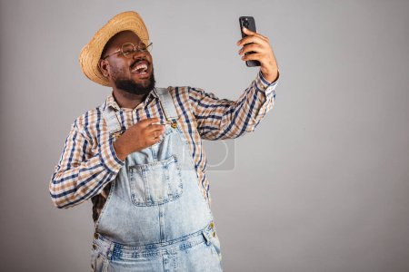 Photo for Brazilian black man wearing country clothes from festa junina,festa de so joo. arrai, holding smartphone, making selfie, video call. - Royalty Free Image