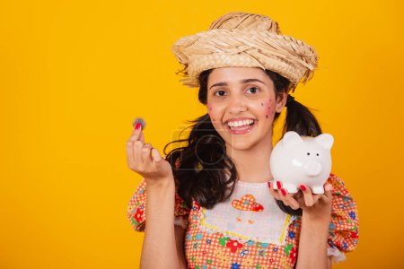 Photo for Beautiful Brazilian woman, with June party clothes, holding coin and piggy bank. - Royalty Free Image