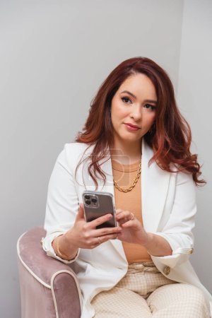 Photo for Brazilian woman, redhead, beautician, beauty professional, sitting in a chair. holding smartphone - Royalty Free Image