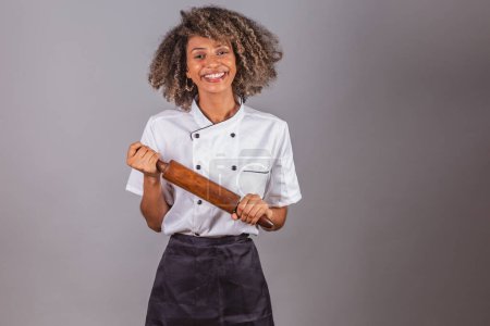 Photo for Young black Brazilian woman, cook, masterchef, wearing restaurant uniform. holding wooden rolling pin for preparing pasta, bread and pizza. - Royalty Free Image