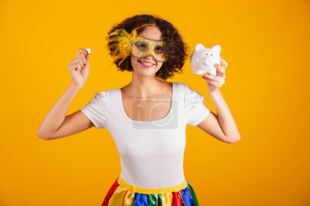 Photo for Beautiful Brazilian woman, dressed in carnival clothes, colorful skirt and white shirt. wearing mascara, holding piggy bank and coin. - Royalty Free Image