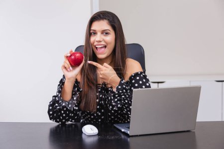 Photo for Beautiful Brazilian woman, entrepreneur, nutritionist, holding an apple, sitting at a table with a notebook. Home office. Good habits. - Royalty Free Image