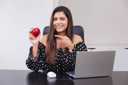 Photo for Beautiful Brazilian woman, entrepreneur, nutritionist, holding an apple, sitting at a table with a notebook. Home office. Good habits. - Royalty Free Image