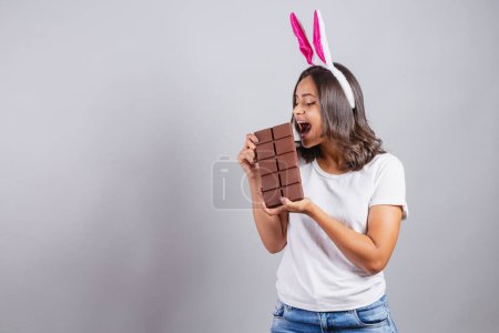 Foto de Mujer brasileña, con orejas de conejo de Pascua, sonriendo felizmente, sosteniendo la barra de chocolate de Pascua. - Imagen libre de derechos