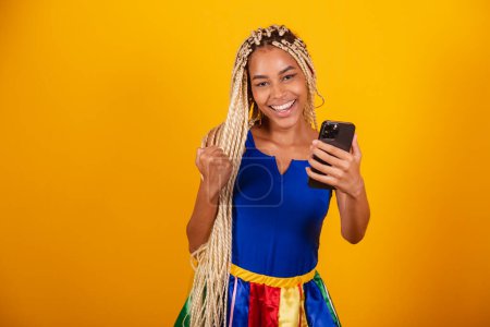 Foto de Hermosa mujer negra, brasileña con trenzas, con ropa para el carnaval. Sostiene el smartphone. aplausos. - Imagen libre de derechos