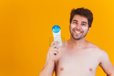 Foto de Hombre guapo con barba de vacaciones con traje de baño sosteniendo la botella de loción protector solar mirando positiva y feliz de pie y sonriendo con una sonrisa confiada mostrando los dientes - Imagen libre de derechos