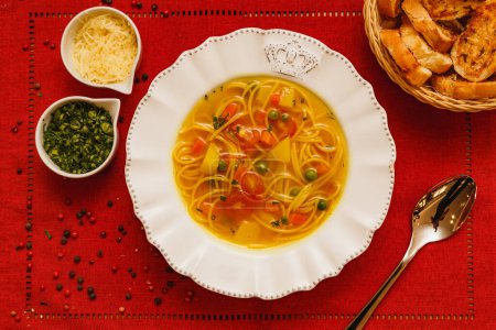 Foto de Deliciosa sopa de fideos con verduras, sopa de fideos con verduras, sobre un fondo de madera, fondo de tela roja, con tostadas, perejil y parmesano, alimentos brasileños de invierno, cocina gourmet - Imagen libre de derechos