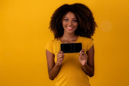 Foto de Mujer afro con el pelo rizado apuntando al teléfono celular sobre fondo amarillo - Imagen libre de derechos