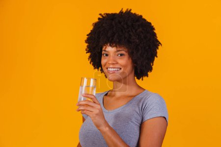 Téléchargez les photos : Jeune femme noire buvant de l'eau en fond jaune. Jeune fille avec verre d'eau - en image libre de droit