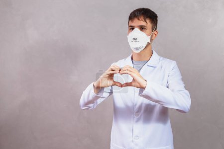 Téléchargez les photos : Jeune médecin portant un masque protecteur faire un cœur avec sa main - en image libre de droit