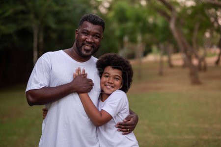 Téléchargez les photos : Père noir et fils dans le parc se tenant souriant regardant caméra - en image libre de droit