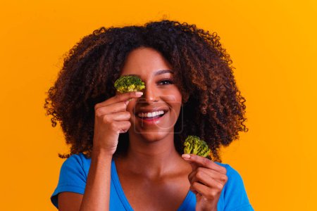 Foto de Mujer afro feliz comiendo brocolis. mujer con brocolis sobre fondo amarillo. concepto de alimentación saludable - Imagen libre de derechos