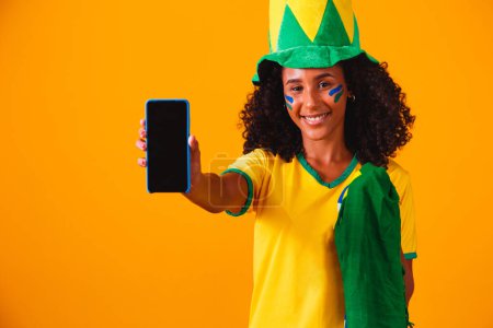 Foto de Abanico brasileño. retrato, ventilador brasileño mostrando su teléfono celular, vestido como un ventilador de fútbol o partido de fútbol sobre fondo amarillo. Colores de Brasil.Copa del Mundo - Imagen libre de derechos