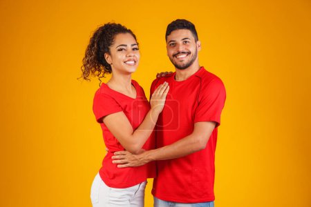 Téléchargez les photos : Portrait de couple de contenu en vêtements de base souriant à la caméra tandis que la femme mettant sa tête sur l'épaule masculine isolé sur fond jaune, il est surpris et heureux - en image libre de droit