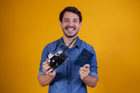 Photo for Smiling handsome tourist man holding a passport and a photo camera on yellow background. - Royalty Free Image