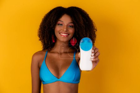 Téléchargez les photos : Femme afro tenant une bouteille de crème solaire sur blanc. Présentation d'un produit. Concept d'été, plage et piscine. - en image libre de droit