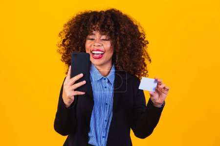 Portrait of excited business woman looking at cell phone while standing and holding credit card isolated over yellow background.