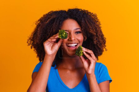 Foto de Mujer afro feliz comiendo brocolis. mujer con brocolis sobre fondo amarillo. concepto de alimentación saludable - Imagen libre de derechos