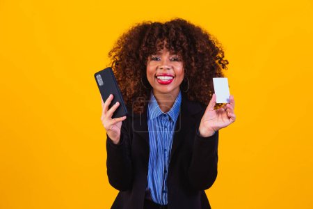 Foto de Retrato de mujer de negocios emocionada mirando el teléfono celular mientras está de pie y sostiene la tarjeta de crédito aislada sobre fondo amarillo. - Imagen libre de derechos