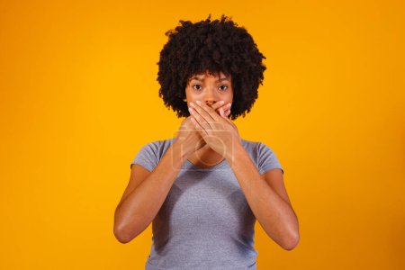 Téléchargez les photos : Femme noire sur fond jaune avec la main dans la bouche, concept de violence, fémicide, racisme et préjugés - en image libre de droit