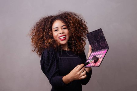 Téléchargez les photos : Afro femme maquilleuse professionnelle tenant pinceaux de maquillage et palette de fard à joues. - en image libre de droit