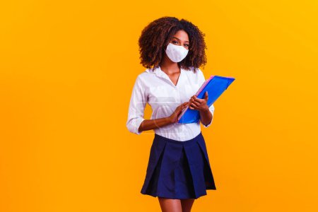 Photo for Afro girl holding books on yellow background wearing protective mask. Return to classes by quarantine - Royalty Free Image