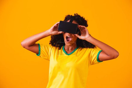 Téléchargez les photos : Un fan brésilien. portrait, fan brésilienne montrant son téléphone portable, habillée en fan de football ou jeu de football sur fond jaune. Couleurs du Brazil.World Cup - en image libre de droit
