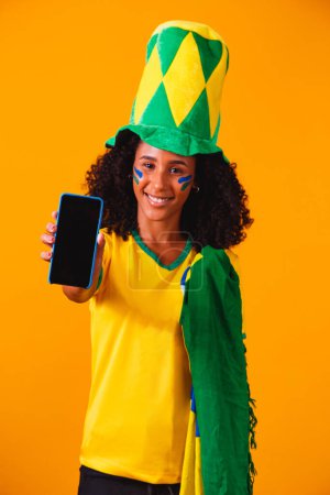 Photo for Brazilian fan. portrait, Brazilian fan showing her cell phone, dressed as a football fan or football game on yellow background. Colors of Brazil.World Cup - Royalty Free Image