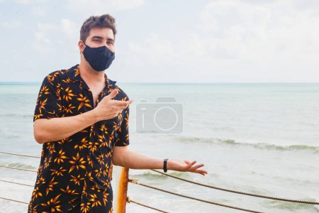 Foto de Primer plano de un joven en la playa usando una máscara para protegerse durante la pandemia señalando - Imagen libre de derechos