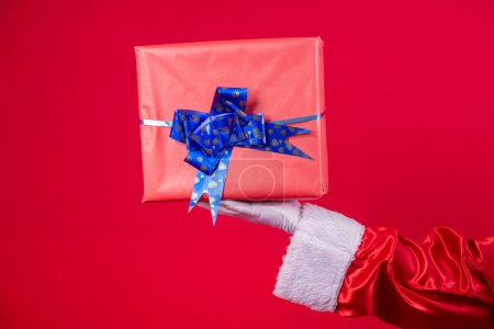 Photo for Santa Claus hand holding Christmas gift box over red background. - Royalty Free Image