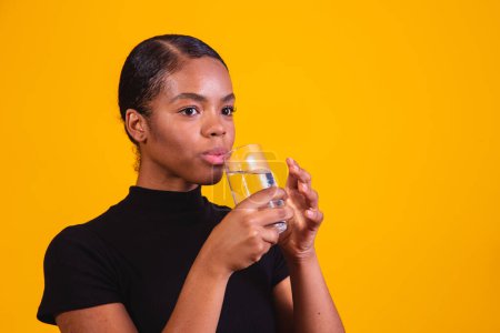 Foto de Mujer afro joven bebiendo agua en fondo amarillo. Chica joven con vaso de agua - Imagen libre de derechos