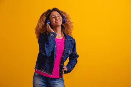 Foto de Hermosa joven con cabello afro escuchando música con sus auriculares sobre fondo amarillo con espacio libre para texto. - Imagen libre de derechos
