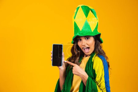 Foto de Abanico brasileño. retrato, ventilador brasileño mostrando su teléfono celular, vestido como un ventilador de fútbol o partido de fútbol sobre fondo amarillo. Colores de Brasil.Copa del Mundo - Imagen libre de derechos