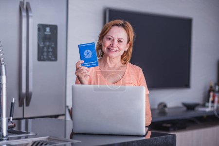Téléchargez les photos : Femme âgée tenant une carte de travail et un ordinateur brésiliens. Femme âgée tenant un cahier d'exercices avec concept de bureau à domicile. - en image libre de droit