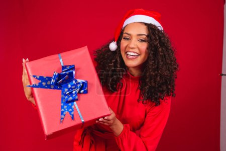 Photo for Portrait of young afro woman in santa claus hat holding a gift on red background. christmas night concept - Royalty Free Image
