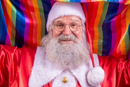 Foto de Santa Claus cubriendo con la bandera del orgullo LGBT. Yo sola. Uno. Mantener el puño en alto, cubriendo la bandera LGBT. Bandera LGBT + sobre fondo rojo. Navidad.. - Imagen libre de derechos