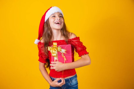 Foto de Linda chica alegre en un sombrero de Navidad en un fondo coloreado sosteniendo un regalo. - Imagen libre de derechos