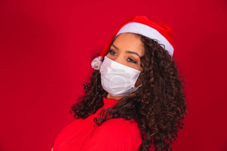 Foto de Joven santa claus mujer en sombrero de Navidad con covid-19 virus coronavirus guardado máscara aislada en el estudio de fondo rojo. Feliz Año Nuevo Celebración Concepto de vacaciones. - Imagen libre de derechos