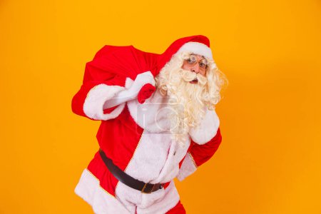 Foto de Santa Claus sobre fondo amarillo sosteniendo bolsa con regalos a sus espaldas. Santa Claus haciendo una sorpresa en la noche de Navidad - Imagen libre de derechos