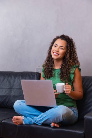 Foto de Sonriendo afro joven mujer usando el ordenador portátil, sentado en el sofá en casa, hermosa chica de compras o chat en línea en la red social, divertirse, ver películas, freelancer trabajando en el proyecto informático - Imagen libre de derechos