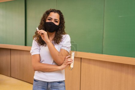 Téléchargez les photos : Jeune professeur avec craie et gomme à effacer à la main portant un masque chirurgical dans la nouvelle normale. - en image libre de droit