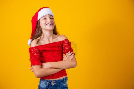 Foto de Retrato de un niño con las manos dobladas, sonriente, listo para ayudar en Nochebuena, con ropa de Navidad y sombrero sobre un fondo amarillo. - Imagen libre de derechos